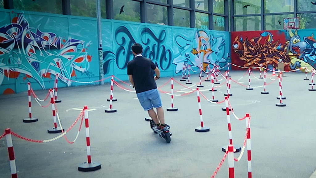 E-Roller Fahrer beim Slalom zwischen rot-weißen Hindernissen. Graffiti-Wand im Hintergrund.