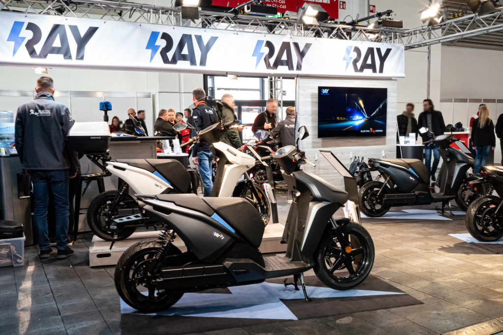 E-Roller der Marke Ray auf einer Messe. Im Hintergrund der Markenname auf einem Banner.